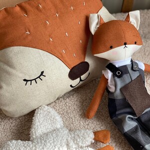 May include: A plush fox toy with a brown and white patterned body, wearing overalls with a black and white plaid pattern. The fox is sitting next to a brown and white fox-shaped pillow with closed eyes.