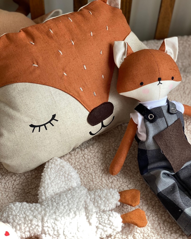 May include: A plush fox toy with a brown and white patterned body, wearing overalls with a black and white plaid pattern. The fox is sitting next to a brown and white fox-shaped pillow with closed eyes.