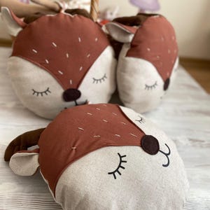May include: Three brown and beige plush pillows shaped like deer heads with closed eyes and stitched details. The pillows are on a light wood surface.