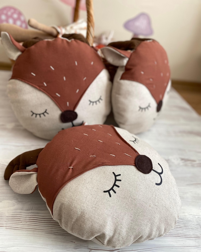 May include: Three brown and beige plush pillows shaped like deer heads with closed eyes and stitched details. The pillows are on a light wood surface.