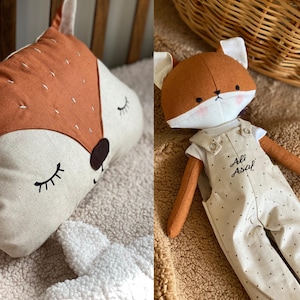 May include: A plush fox toy with a brown and white fur, wearing a beige overall with black polka dots and the text "Ali Asal" embroidered on the front. The toy is sitting on a light brown fuzzy surface.