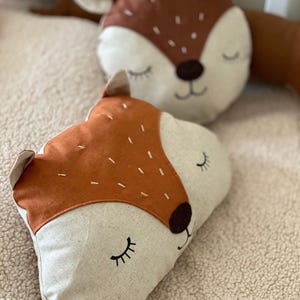 May include: Two brown and white plush pillows with embroidered animal faces. The pillows have closed eyes and a stitched smile. The pillows are on a white fluffy surface.