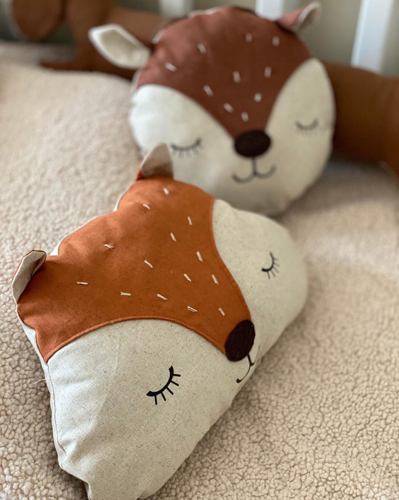 May include: Two brown and white plush pillows with embroidered animal faces. The pillows have closed eyes and a stitched smile. The pillows are on a white fluffy surface.