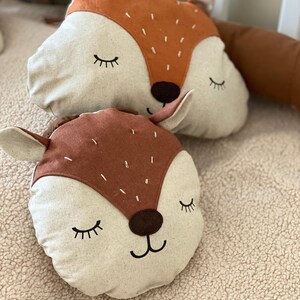 May include: Two plush fox-shaped pillows with brown and beige fabric. The pillows have embroidered eyes, noses, and mouths. The pillows are lying on a beige fuzzy surface.
