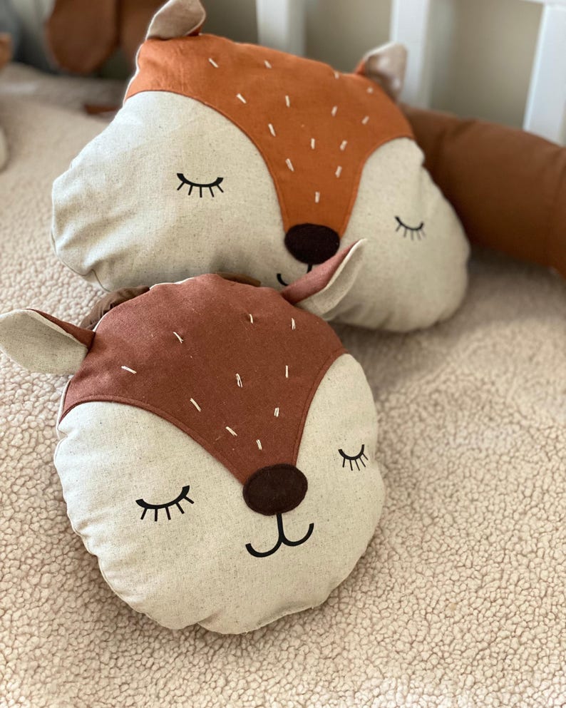 May include: Two plush fox-shaped pillows with brown and beige fabric. The pillows have embroidered eyes, noses, and mouths. The pillows are lying on a beige fuzzy surface.