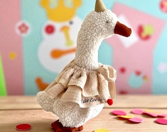 May include: A plush white goose toy wearing a party hat with a red pom-pom and a beige ruffled dress. The goose has a brown beak and feet, with the text "Edie Everly" on the dress. Confetti is scattered around the base.