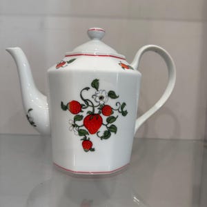 May include: White ceramic teapot with a red strawberry design. The teapot features a curved spout, a rounded handle, and a lid with a small knob. The strawberry design includes red strawberries, green leaves, and white flowers.