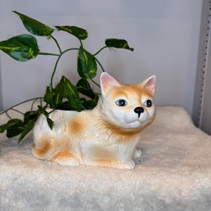 May include: A ceramic cat-shaped planter in cream and orange with green and yellow leafy vines. The cat has blue eyes and a black nose. The planter is sitting on a white, fluffy surface. A decorative item for home decor.
