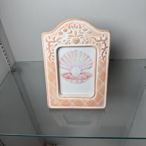 Puede incluir: Un marco de fotos rectangular de cerámica con un acabado en color melocotón claro y un patrón de celosía blanco. El borde superior es arqueado y presenta detalles en relieve de flores y corazones. Contiene una ilustración de una concha.