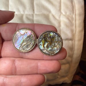 May include: Two circular buttons featuring iridescent abalone shell inlays with swirling patterns of pink, green, and gold. Each button is framed by a thin, textured metallic border with a beaded edge, creating a distinct contrast between the shell.