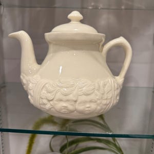 May include: Cream-colored ceramic teapot with a lid and handle. The teapot features a decorative relief of cherubic faces around the body. The spout is curved, and the handle is arched. The teapot is displayed on a glass shelf.