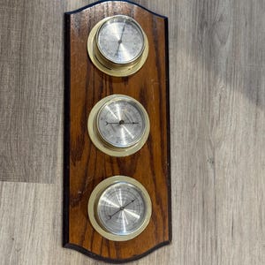 May include: A wooden weather station with three circular gauges. Each gauge has a silver face with black markings and a gold-colored rim. The wooden backing is dark brown with a black border.