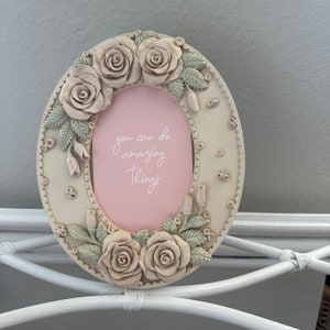 May include: Oval-shaped picture frame with a white background and a pink insert. The frame is decorated with raised white roses and green leaves. The pink insert has the text "you can do amazing things" written in white.