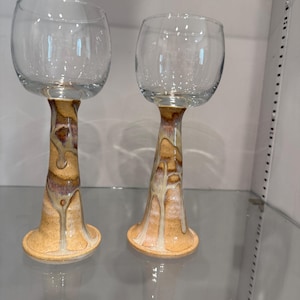 May include: Two clear wine glasses with unique, hand-painted stems. The stems feature a blend of tan, brown, and cream colors, creating a marbled effect. The bases are a solid tan color. The glasses are displayed on a glass shelf.