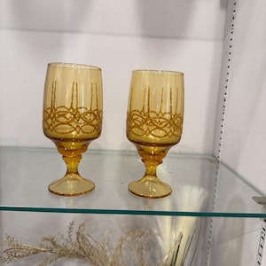 Puede incluir: Dos copas de vino de color ámbar con intrincados diseños dorados. Las copas tienen forma de cuenco redondeado y se asientan sobre una base de pedestal. Se exhiben en una estantería de cristal, lo que sugiere un tema vintage o decorativo.