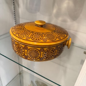 Puede incluir: Una fuente redonda vintage con tapa, de color amarillo cálido. La fuente presenta un patrón repetitivo de diseños circulares marrones. Tiene dos asas pequeñas y un pomo redondo en la tapa. La fuente se exhibe en una estantería de cristal.