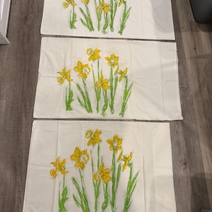 May include: Three rectangular off-white fabric pillowcases, each featuring an identical printed illustration of yellow daffodils with green stems and leaves. The floral design is centered on each piece, showcasing multiple blooming flowers.
