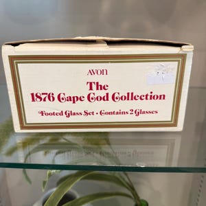 Puede incluir: Una taza de cerámica blanca con un asa negra y el texto "But First, Coffee" en negrita negra. La taza se encuentra sobre un fondo borroso, sugiriendo un ambiente acogedor.