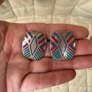 Op de afbeelding: Een paar rechthoekige oorbellen met afgeronde hoeken, voorzien van een zilverkleurige metalen basis ingelegd met geometrische emaille patronen in paars, rood, blauwgroen en oranje. Het ontwerp toont een ruitvorm omringd door schuine strepen.