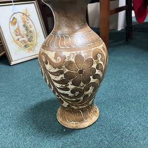 Puede incluir: Un jarrón de cerámica marrón con un diseño floral blanco. El jarrón tiene una base ancha y un cuello estrecho.