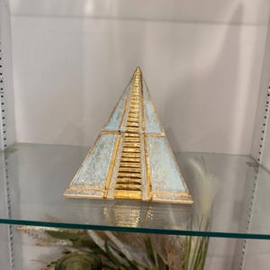 Può includere: Una scultura decorativa a forma di piramide, nei toni dell'azzurro e dell'oro. La piramide presenta un design a gradini con dettagli dorati e una superficie strutturata. È esposta su una mensola di vetro.