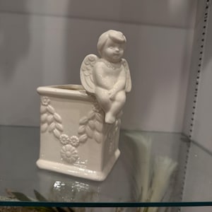 May include: A cream-colored ceramic planter with a square base and floral relief detailing. A small cherub figure with wings sits on the planter's edge. The planter is displayed on a glass shelf.