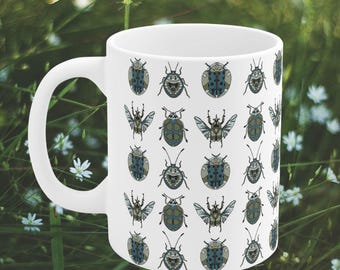Puede incluir: Taza de cerámica blanca con un patrón repetido de escarabajos azules y verdes. Los escarabajos son estilizados y tienen alas, patas y antenas.