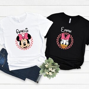 以下が含まれることがあります： 2枚のTシャツ。1枚は白、もう1枚は黒で、漫画のキャラクターのデザインが施されています。白いシャツには、名前「Amelia」が上に書かれたミニーマウスのグラフィックがあります。黒いシャツには、名前「Emma」が上に書かれたデイジーダックのグラフィックがあります。