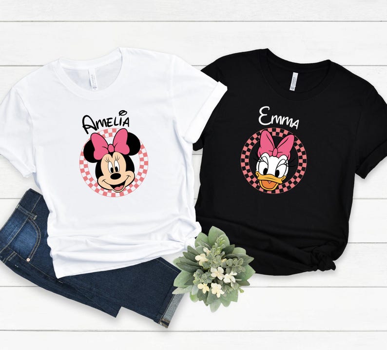 以下が含まれることがあります： 2枚のTシャツ。1枚は白、もう1枚は黒で、漫画のキャラクターのデザインが施されています。白いシャツには、名前「Amelia」が上に書かれたミニーマウスのグラフィックがあります。黒いシャツには、名前「Emma」が上に書かれたデイジーダックのグラフィックがあります。