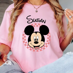 以下が含まれることがあります： ピンク色のTシャツ。ミッキーマウスのグラフィックと「Susan」という名前が黒字でプリントされています。ミッキーマウスのデザインは、ピンクと白のチェック柄の円の中に配置され、遊び心のあるパーソナライズされた外観になっています。