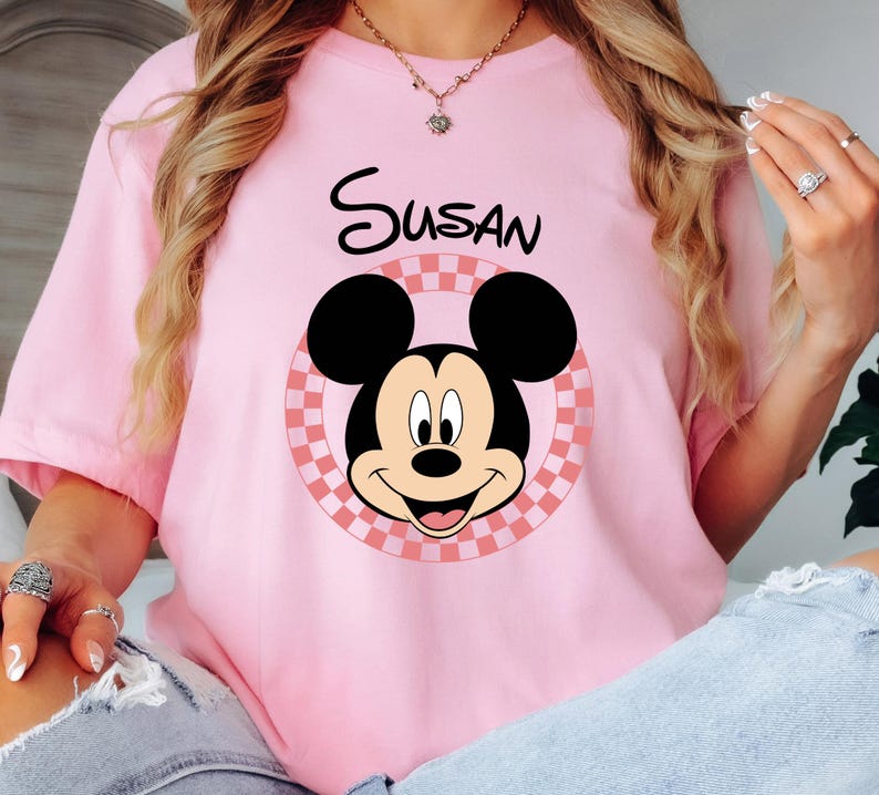 以下が含まれることがあります： ピンク色のTシャツ。ミッキーマウスのグラフィックと「Susan」という名前が黒字でプリントされています。ミッキーマウスのデザインは、ピンクと白のチェック柄の円の中に配置され、遊び心のあるパーソナライズされた外観になっています。