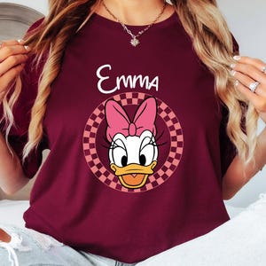 以下が含まれることがあります： バーガンディ色のTシャツ。デイジーダックの漫画のグラフィック、ピンクのリボン、市松模様の円のデザインが特徴です。「Emma」という名前がグラフィックの上に白で印刷されています。Tシャツは単色です。