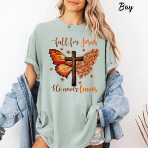 May include: A light sage green t-shirt featuring a graphic print of a wooden cross centered between an orange butterfly wing and a cluster of autumn leaves. The text reads "fall for Jesus" above and "He never leaves" below the graphic.