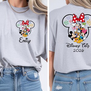a woman wearing a gray t-shirt with a Disney-themed design featuring Minnie Mouse and other Disney characters. The t-shirt has a personalized name printed on it, and the back of the shirt displays the text "Disney Trip 2026".