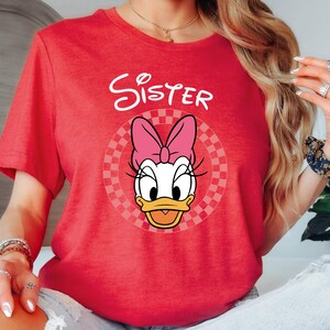 以下が含まれることがあります： 赤いTシャツ。デイジーダックのグラフィック、ピンクのリボン、白いスクリプトで「Sister」の文字が描かれています。グラフィックはチェック柄の円の中に配置されています。Tシャツは半袖でクルーネックです。