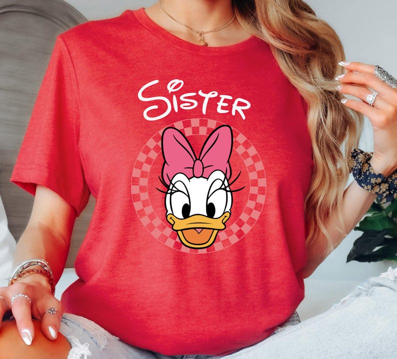 以下が含まれることがあります： 赤いTシャツ。デイジーダックのグラフィック、ピンクのリボン、白いスクリプトで「Sister」の文字が描かれています。グラフィックはチェック柄の円の中に配置されています。Tシャツは半袖でクルーネックです。