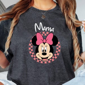 以下が含まれることがあります： 「ママ」の白文字がプリントされた濃いグレーのTシャツ。ミニーマウスのグラフィックは、ピンクのリボン、黒い耳、ピンクと白のチェック柄の円で構成されています。