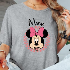 以下が含まれることがあります： ミニーマウスのグラフィックが特徴のライトグレーのTシャツ。グラフィックには、ピンクのリボン、黒い耳、黒い筆記体で書かれた「ママ」という文字が含まれています。ミニーマウスの画像は、ピンクと白のチェック柄の背景に配置されています。