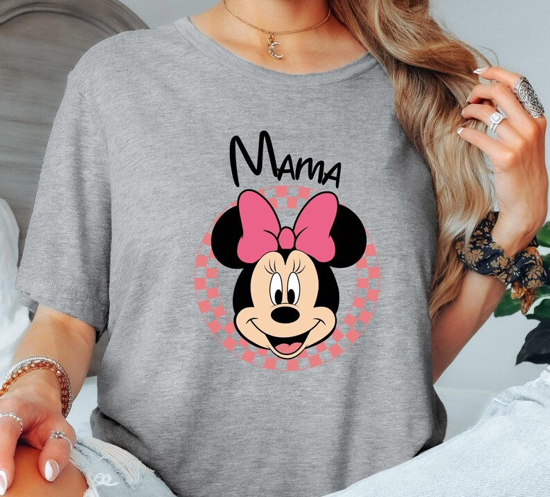 以下が含まれることがあります： ミニーマウスのグラフィックが特徴のライトグレーのTシャツ。グラフィックには、ピンクのリボン、黒い耳、黒い筆記体で書かれた「ママ」という文字が含まれています。ミニーマウスの画像は、ピンクと白のチェック柄の背景に配置されています。