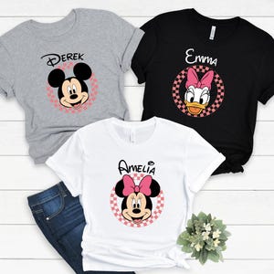 以下が含まれることがあります： 漫画のキャラクターデザインが施された半袖Tシャツ3枚。グレーのシャツには「デレク」という名前のミッキーマウスが描かれています。黒いシャツには「エマ」という名前のデイジーダックが描かれています。白いシャツには「アメリア」という名前のミニーマウスが表示されています。