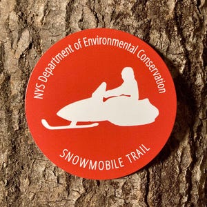 May include: A round, red sticker with white text and a white graphic of a person riding a snowmobile. The text reads "NYS Department of Environmental Conservation" and "Snowmobile Trail". The sticker is on a brown, textured surface.