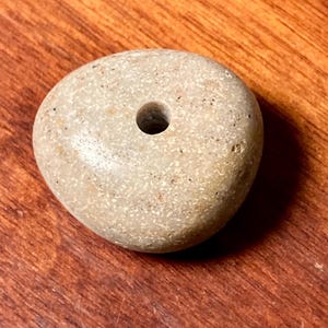 May include: A rounded, light grey stone with a hole through the middle. The stone has a speckled texture. The background is a wooden surface.