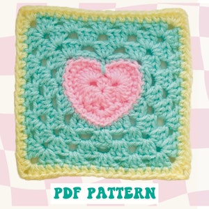 May include: A crochet granny square with a pink heart in the center. The square is bordered with a yellow yarn and the heart is made with a pink yarn.