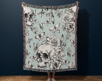 May include: A woven throw blanket with a light blue background featuring a black and white skull design with floral accents and butterflies.