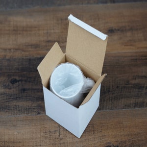 May include: A white ceramic mug is inside a cardboard box with a white lining. The mug is wrapped in plastic.