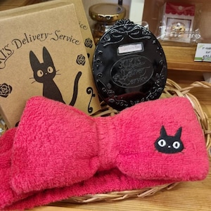 May include: A red headband with a black cat design and a bow. The headband is in a wicker basket with a black compact mirror with a cat design and the text "Kiki's Delivery Service" on it. There is a brown box with the text "Kiki's Delivery Service" and a black cat design on it.
