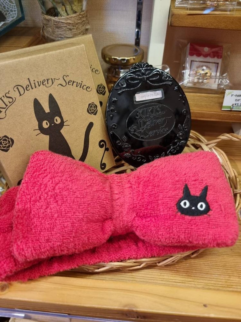 May include: A red headband with a black cat design and a bow. The headband is in a wicker basket with a black compact mirror with a cat design and the text "Kiki's Delivery Service" on it. There is a brown box with the text "Kiki's Delivery Service" and a black cat design on it.
