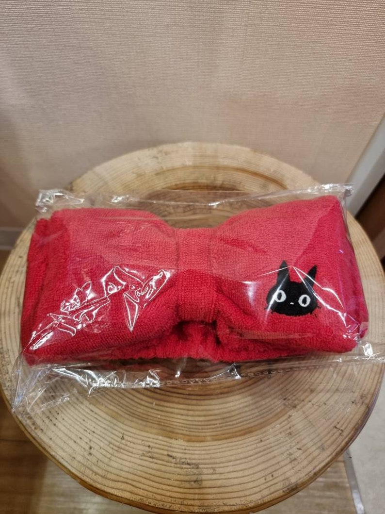 May include: Red headband with a black cat design. The headband is made of a soft, plush material and is in a bow shape.