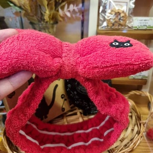 May include: Red terry cloth headband with a black cat design and white stripes. The headband is folded in half and is sitting on a wicker basket.