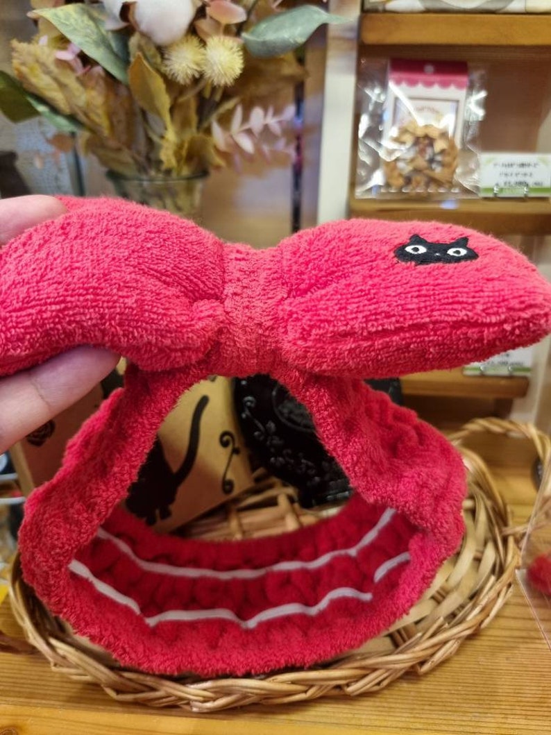 May include: Red terry cloth headband with a black cat design and white stripes. The headband is folded in half and is sitting on a wicker basket.